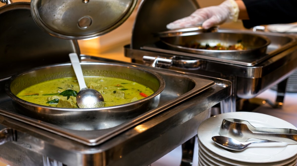 A buffet tray of green Thai curry with its own clean serving spoon, a gloved hand lifting the lid, other dishes and clean utensils set apart