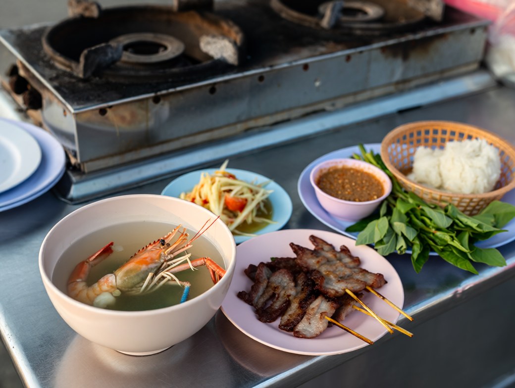 A Thai street-food spread of tom yum soup, green papaya salad, grilled pork skewers, sticky rice and fresh herbs on a stall counter