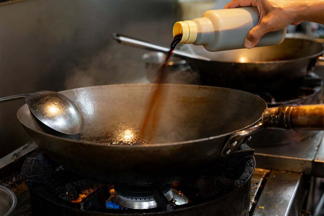 A cook pouring dark soy sauce into a wok on a gas burner in a busy Thai kitchen, steam rising, a ladle on the rim