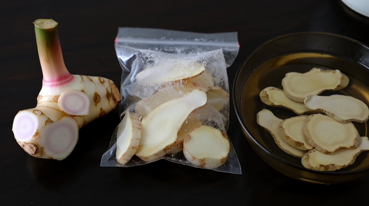Three forms of galangal in a row, a fresh knob with pink-ringed coins, frozen slices in a sealed bag, and slices softening in a bowl of water
