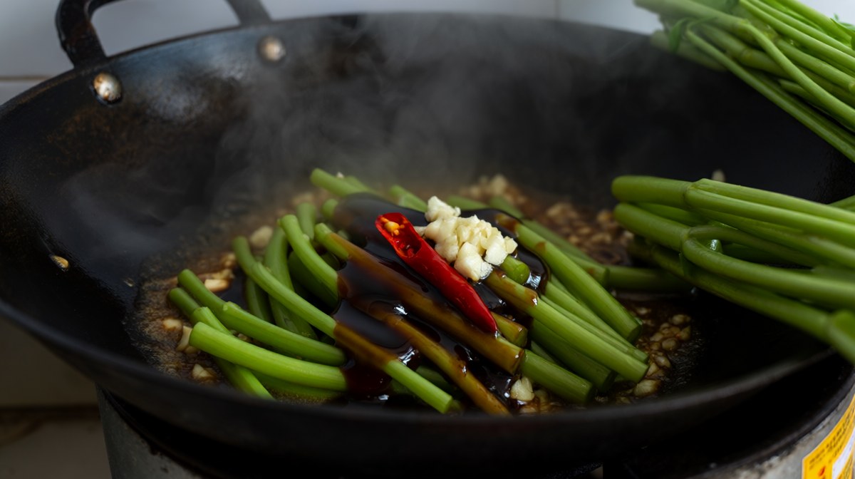 Water spinach stir-frying in a smoking wok with garlic and chilli, steam rising, a fresh bunch beside it