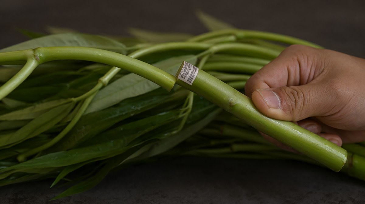 A hand bending a water spinach stem about to snap it, beside a full fresh bunch on a dark surface