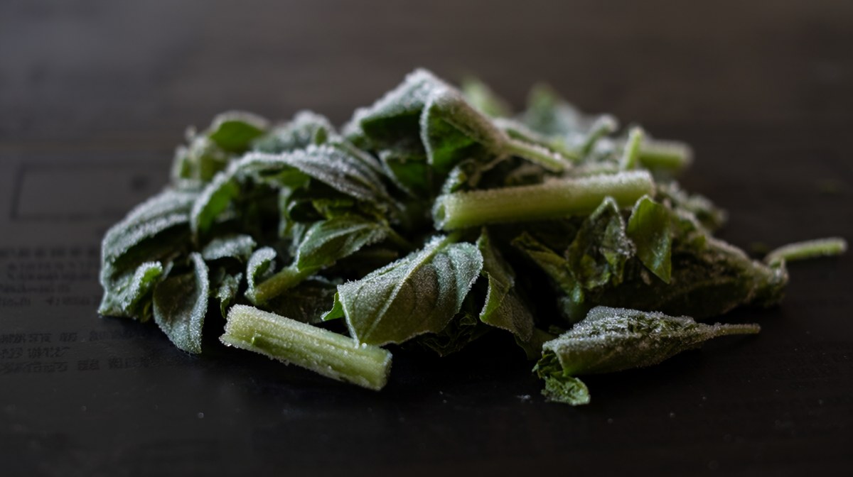A pile of loose frozen Thai basil leaves and chopped stems, frosted and separated on a dark surface
