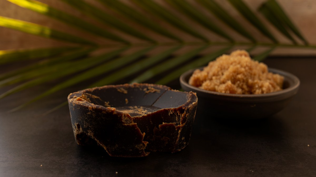 A broken block of dark palm sugar with a palm frond behind it and a bowl of soft brown sugar beside it
