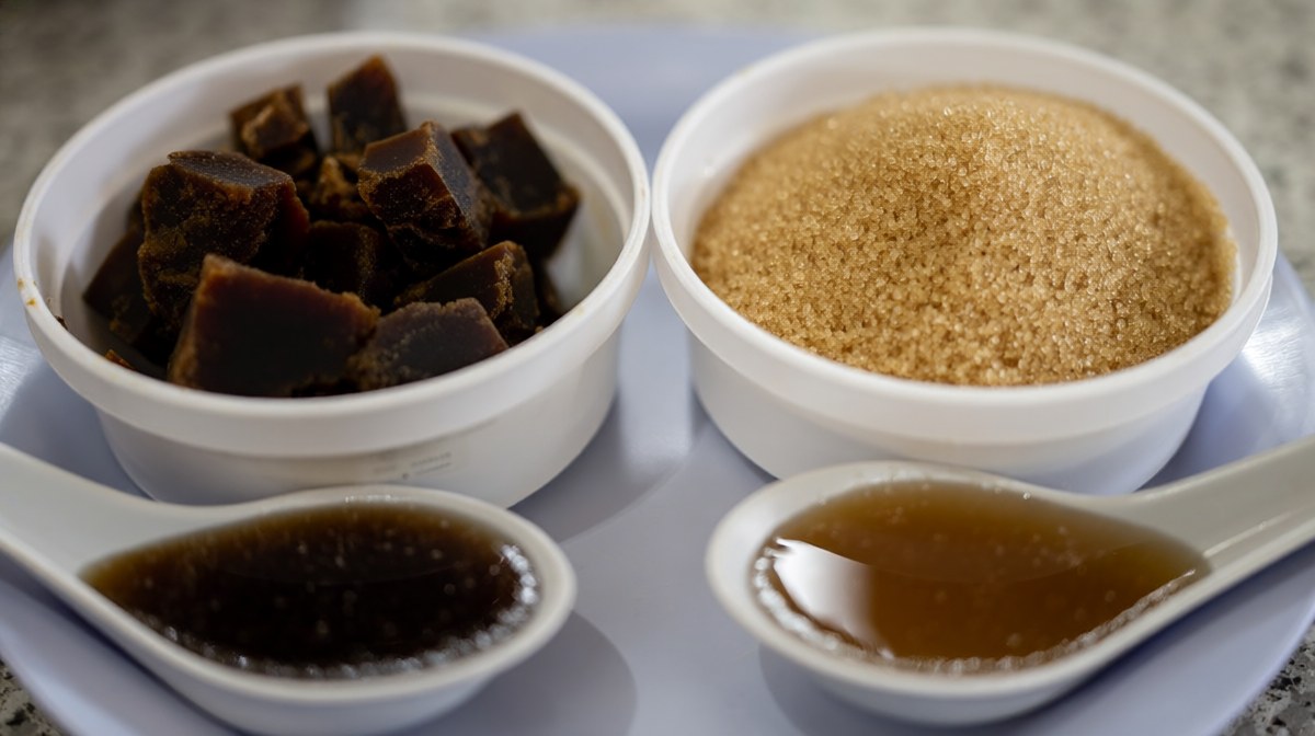 Two bowls side by side of dark palm sugar chunks and fine light brown sugar, with melted spoons below