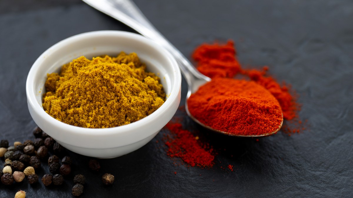 A bowl of coarse yellow curry powder with whole spices scattered beside it and a second red powder