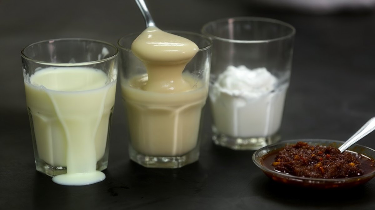 Three glasses in a row of thin pale evaporated milk, thick condensed milk and white coconut cream beside a dish of dark roasted chilli jam