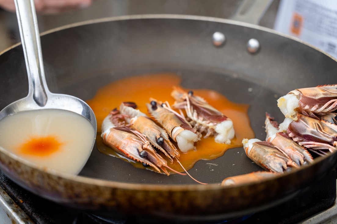 Prawn heads pressed in a hot pan with orange fat pooling out, and a faint orange bloom spreading across a ladle of pale broth beside it