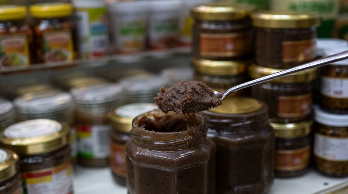 A small jar of dark shrimp paste with a spoon lifting some out, on a shelf of Thai pantry items