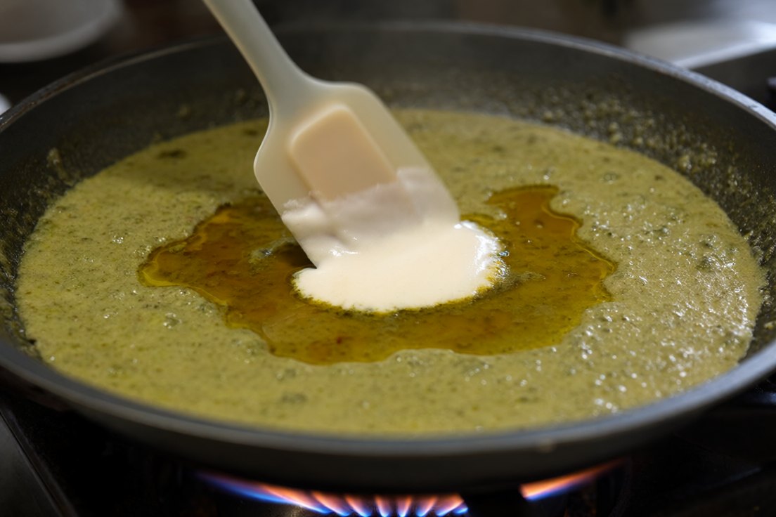 A spoon adding thick coconut cream into the centre of a grainy split green curry while a spatula stirs it in slow small circles on the lowest heat