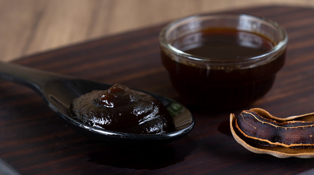 A spoonful of dark tamarind paste beside a glass of concentrate and cracked tamarind pods